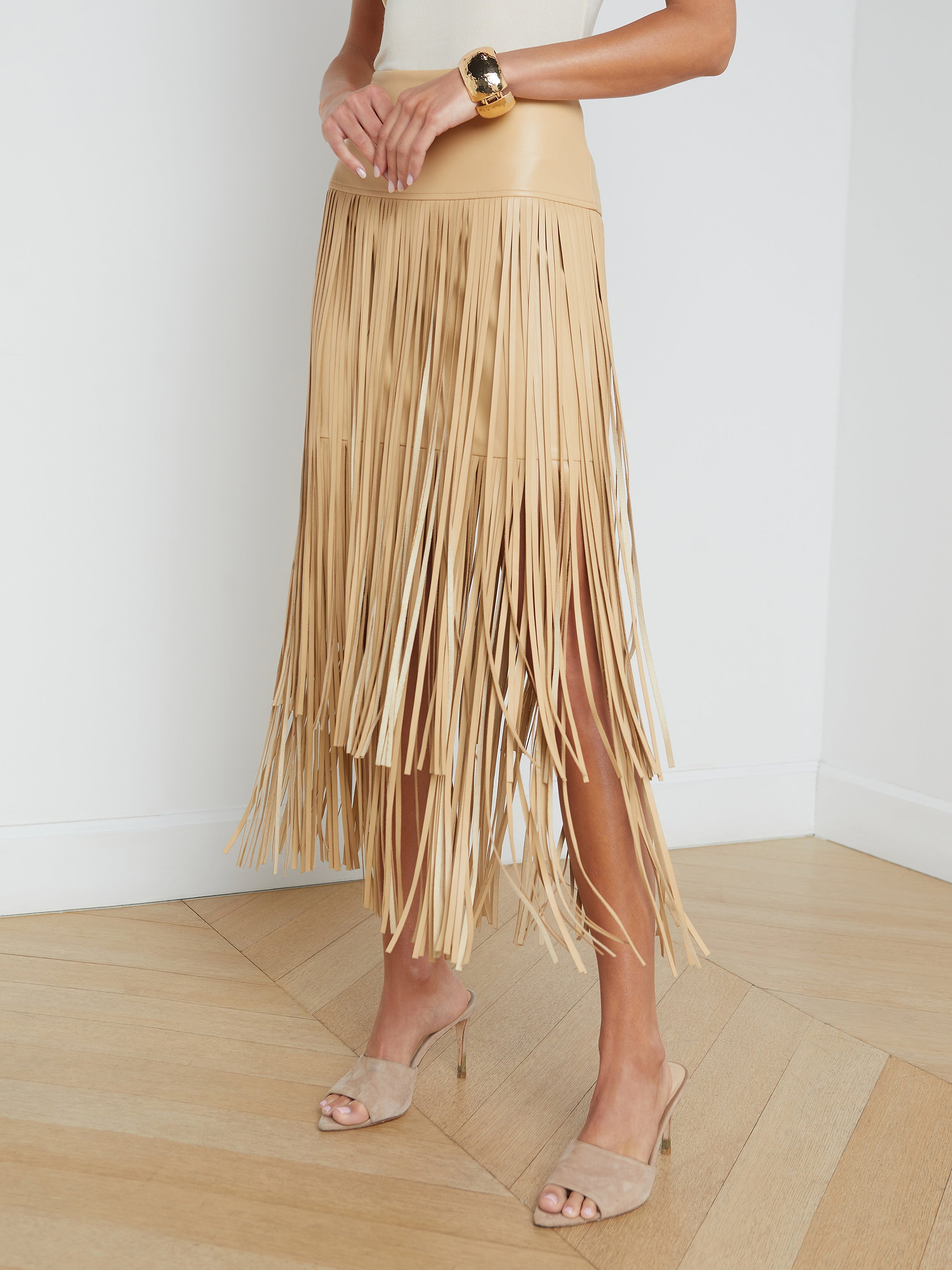 A person wearing the Karolina Fringe Skirt, a white top, beige heeled sandals, and a gold bracelet stands against a plain white background.