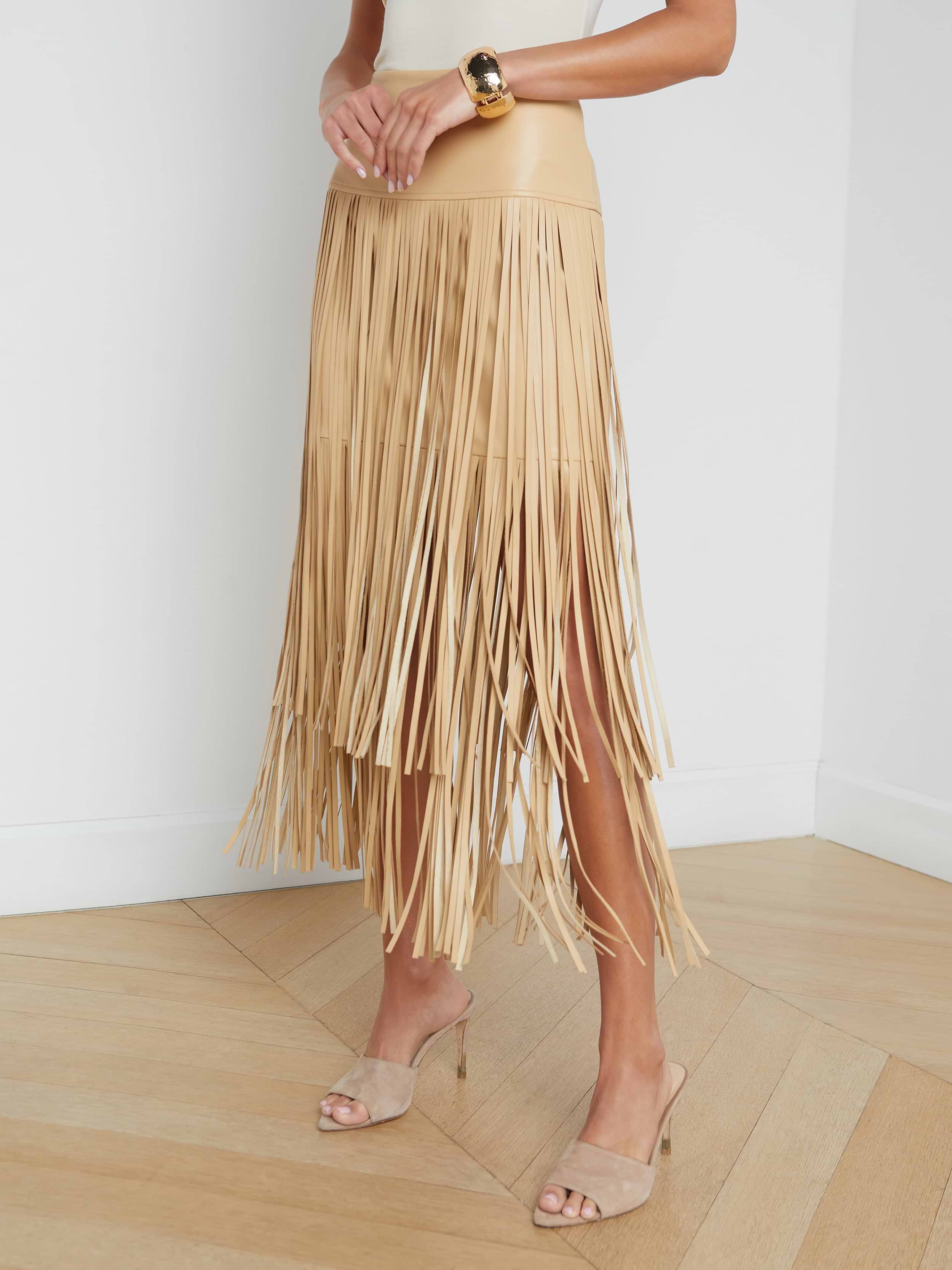 A person wearing the Karolina Fringe Skirt, a white top, beige heeled sandals, and a gold bracelet stands against a plain white background.