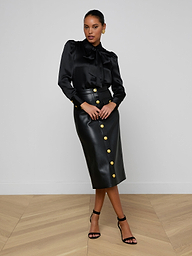 A woman poses in a black long-sleeve blouse with a bow and the Milann Vegan Leather Skirt, a black midi skirt adorned with gold buttons, paired with black heels against a plain white background.