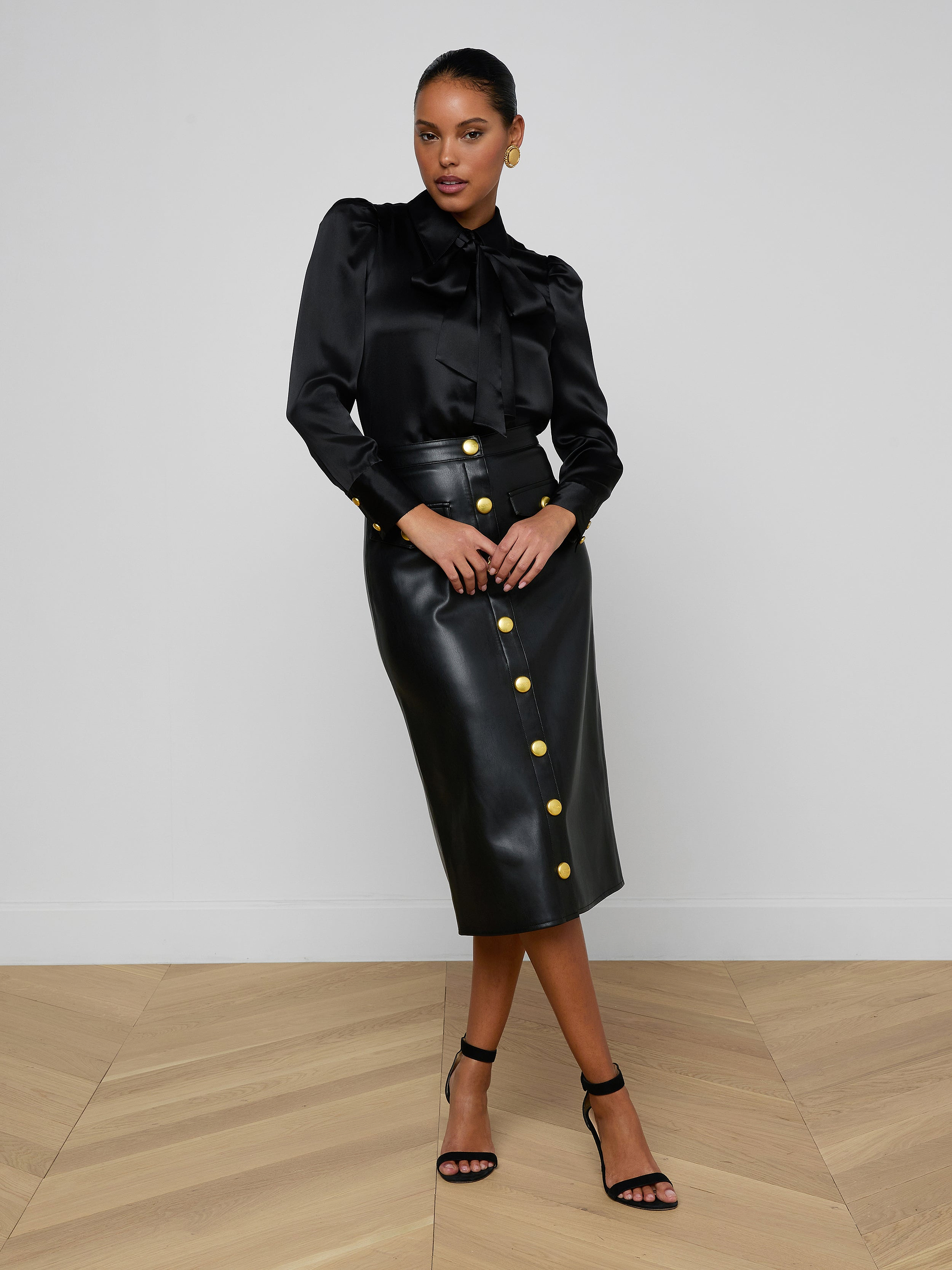 A woman poses in a black long-sleeve blouse with a bow and the Milann Vegan Leather Skirt, a black midi skirt adorned with gold buttons, paired with black heels against a plain white background.