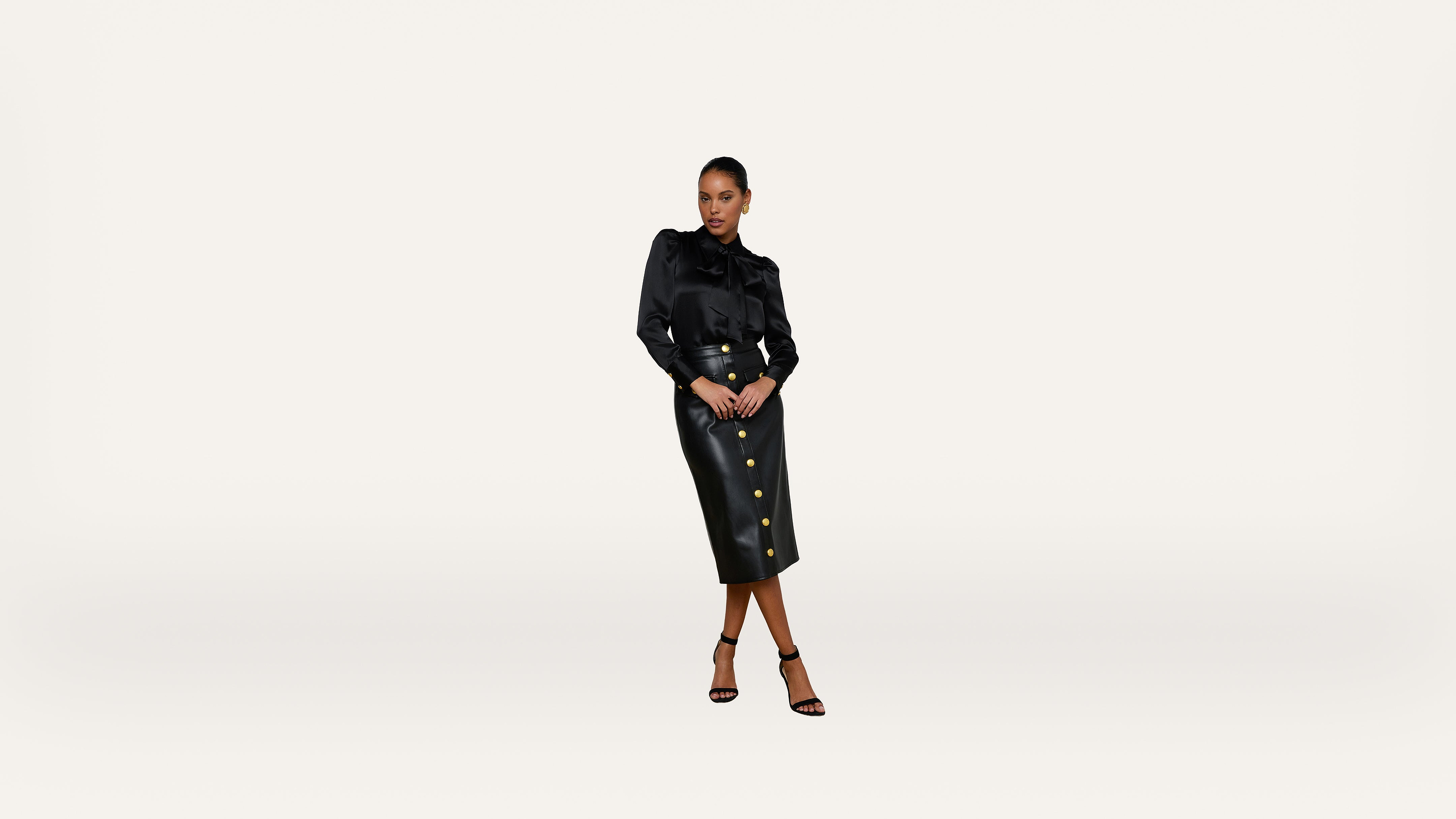 A woman stands confidently in a black blouse and a high-waisted black leather skirt with gold buttons, wearing black heeled sandals, on a plain white background.
