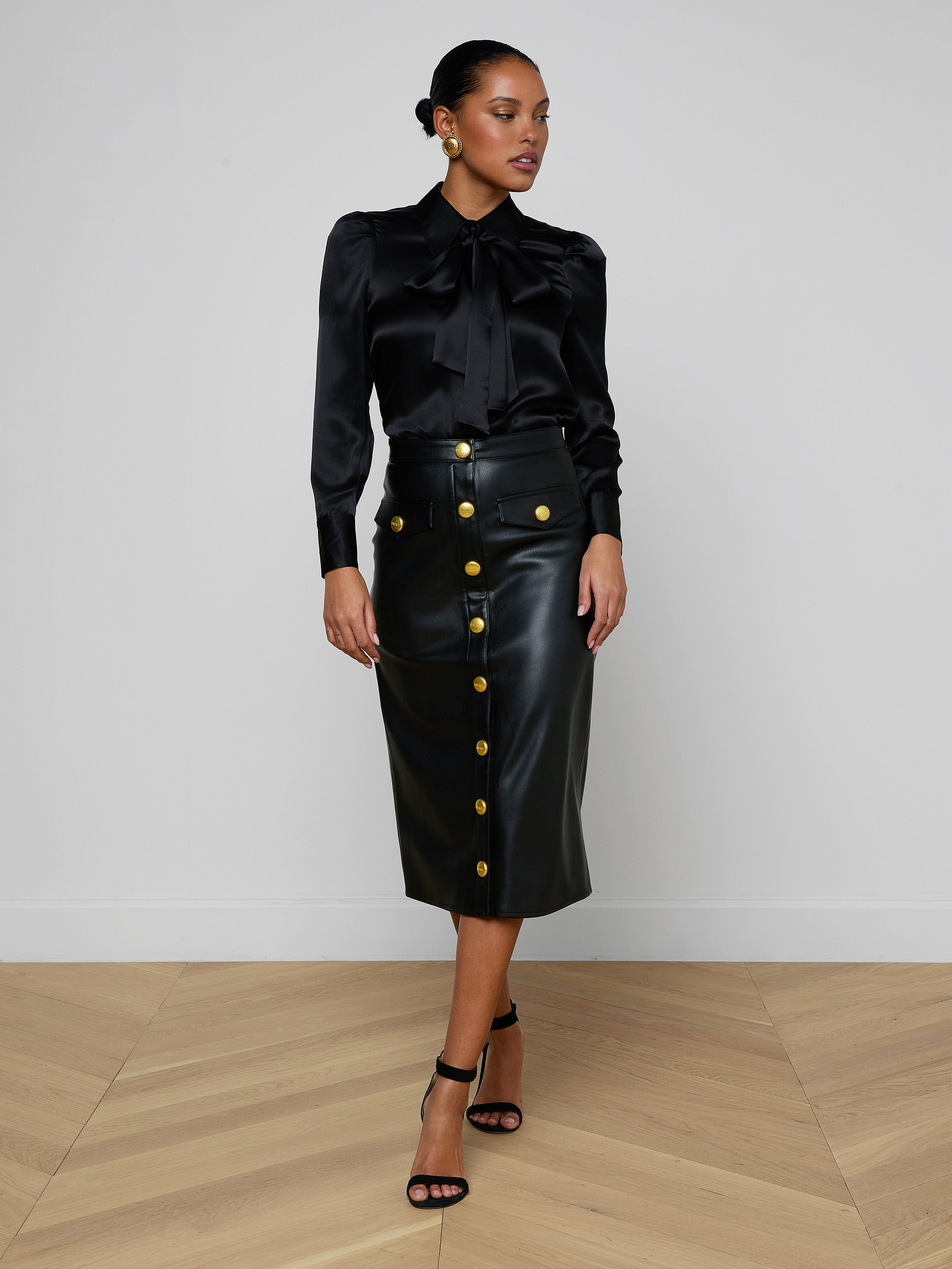 A woman stands confidently against a plain light background, wearing a black satin blouse paired with the high-waisted Milann Vegan Leather Skirt, which features gold buttons, and finishes the look with black heeled sandals.