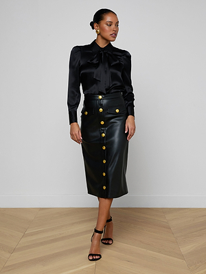 A woman stands indoors wearing a black satin blouse, the Milann Vegan Leather Skirt with gold buttons and a midi hem, black heeled sandals, and gold earrings. The background is plain and neutral.