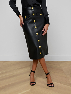 A woman stands on a wooden floor wearing the black, high-waisted Milann Vegan Leather Skirt with large gold buttons and a midi hem, paired with a black blouse and black heeled sandals. Her arms are relaxed at her sides.