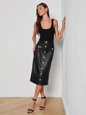 A woman with long brown hair stands indoors against a white wall, wearing the L'AGENCE Milann Vegan Leather Skirt with gold accent buttons, a black sleeveless top, and black heeled sandals, gazing to the side.