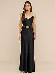 Dressed in the Ryker Silk Maxi Skirt, the woman showcases minimalist sophistication. She pairs the flowing black skirt with a sleeveless top featuring a V-neckline and cinches her waist with a wide black belt adorned by an oversized gold buckle. Her long, wavy brown hair and subtle accessories complete the look as she stands against a plain white background.