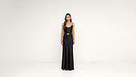 A woman stands facing forward, wearing a long, sleeveless black dress with a gold belt, against a plain white background.