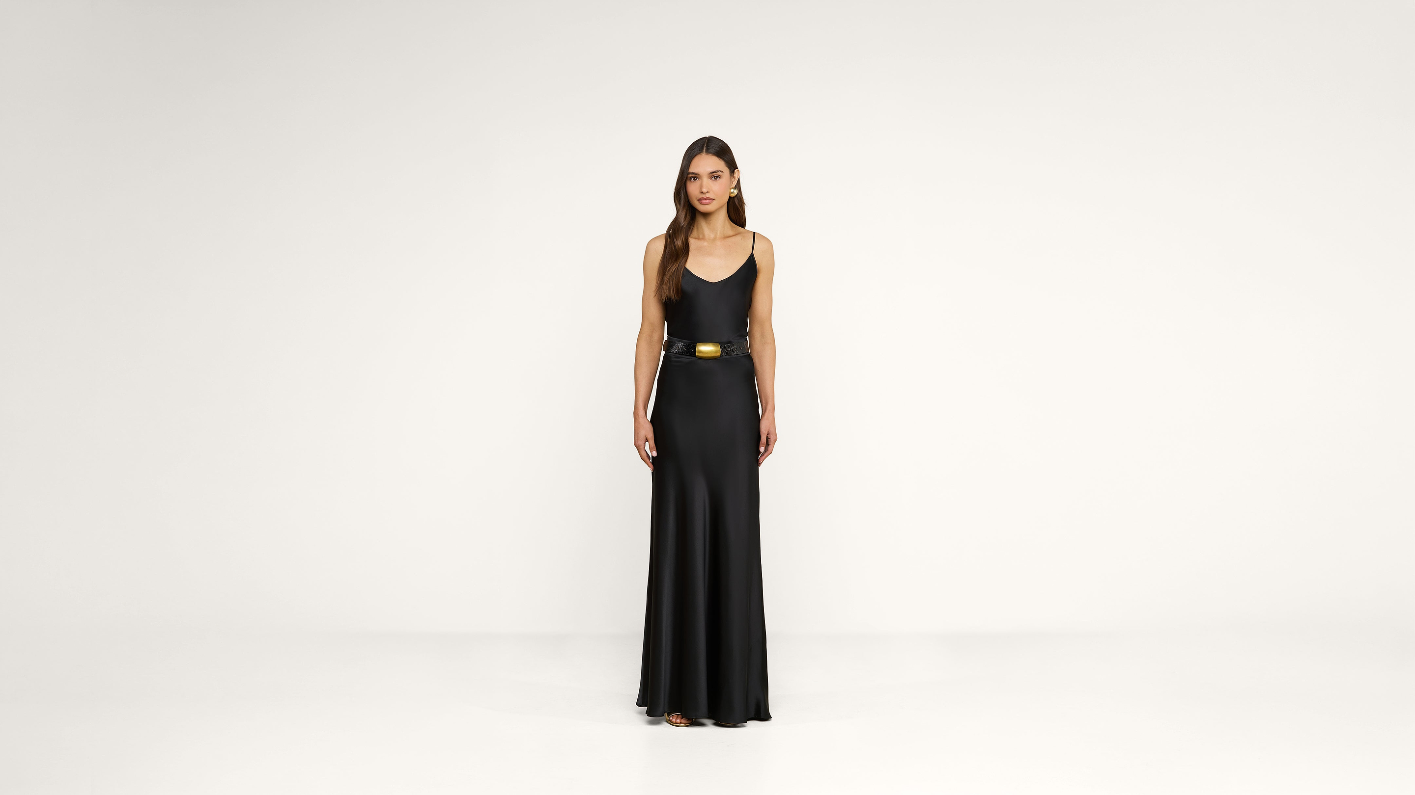 A woman stands facing forward, wearing a long, sleeveless black dress with a gold belt, against a plain white background.