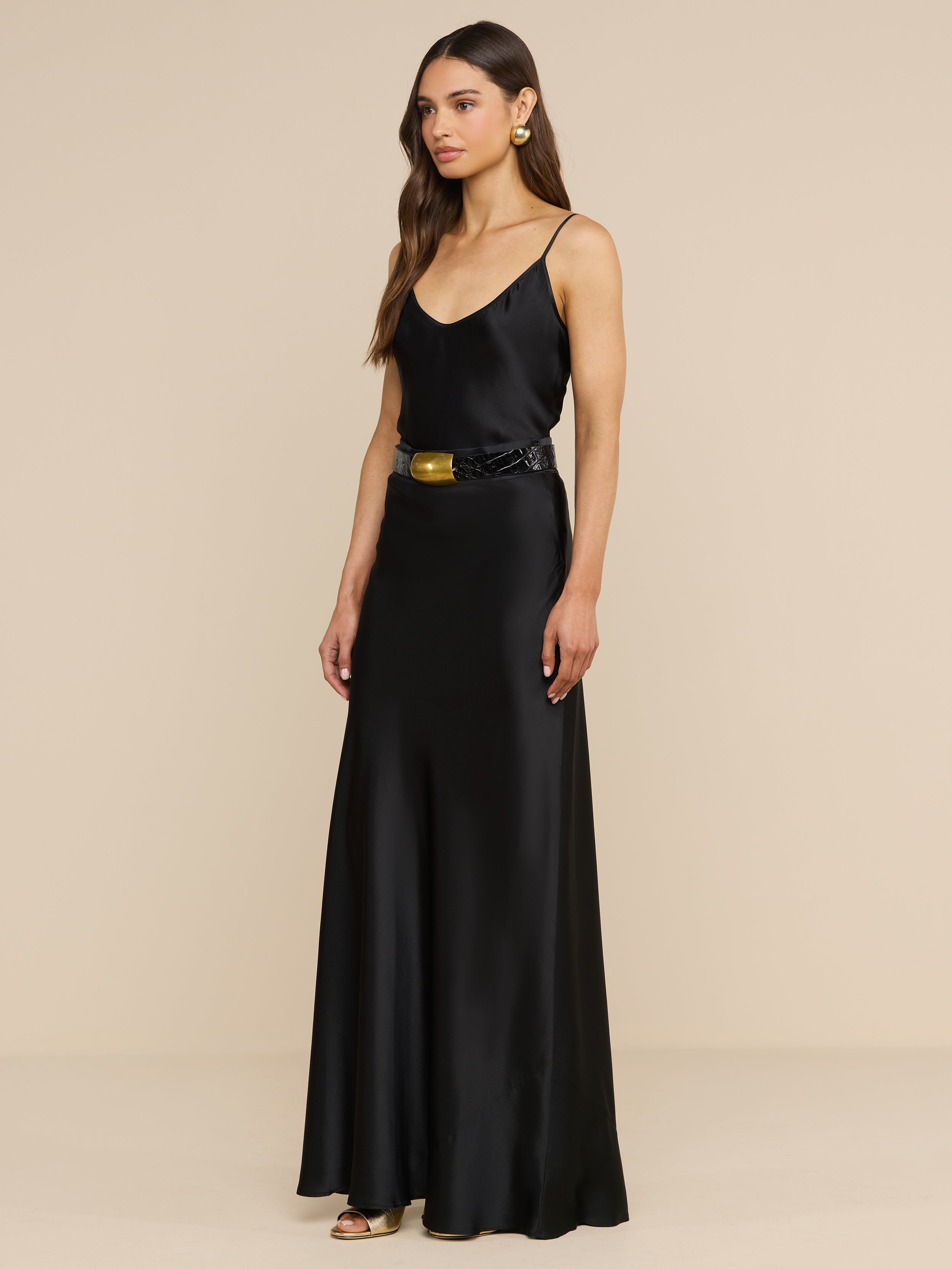 A woman stands in a plain white room, wearing the Ryker Silk Maxi Skirt styled as a long, bias-cut black satin skirt. She pairs it with a sleeveless top featuring thin straps and a black belt with a gold buckle. Gold earrings and open-toed heels complete her look, embodying minimalist sophistication.