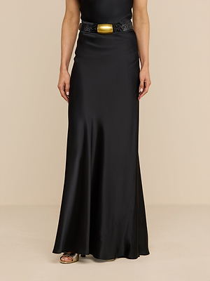 Un mannequin porte la jupe longue « Ryker Silk » en noir, associée à une ceinture noire ornée dune boucle dorée imposante. Le bas du torse et les jambes se détachent sur un fond beige uni, soulignant ainsi une sophistication minimaliste.