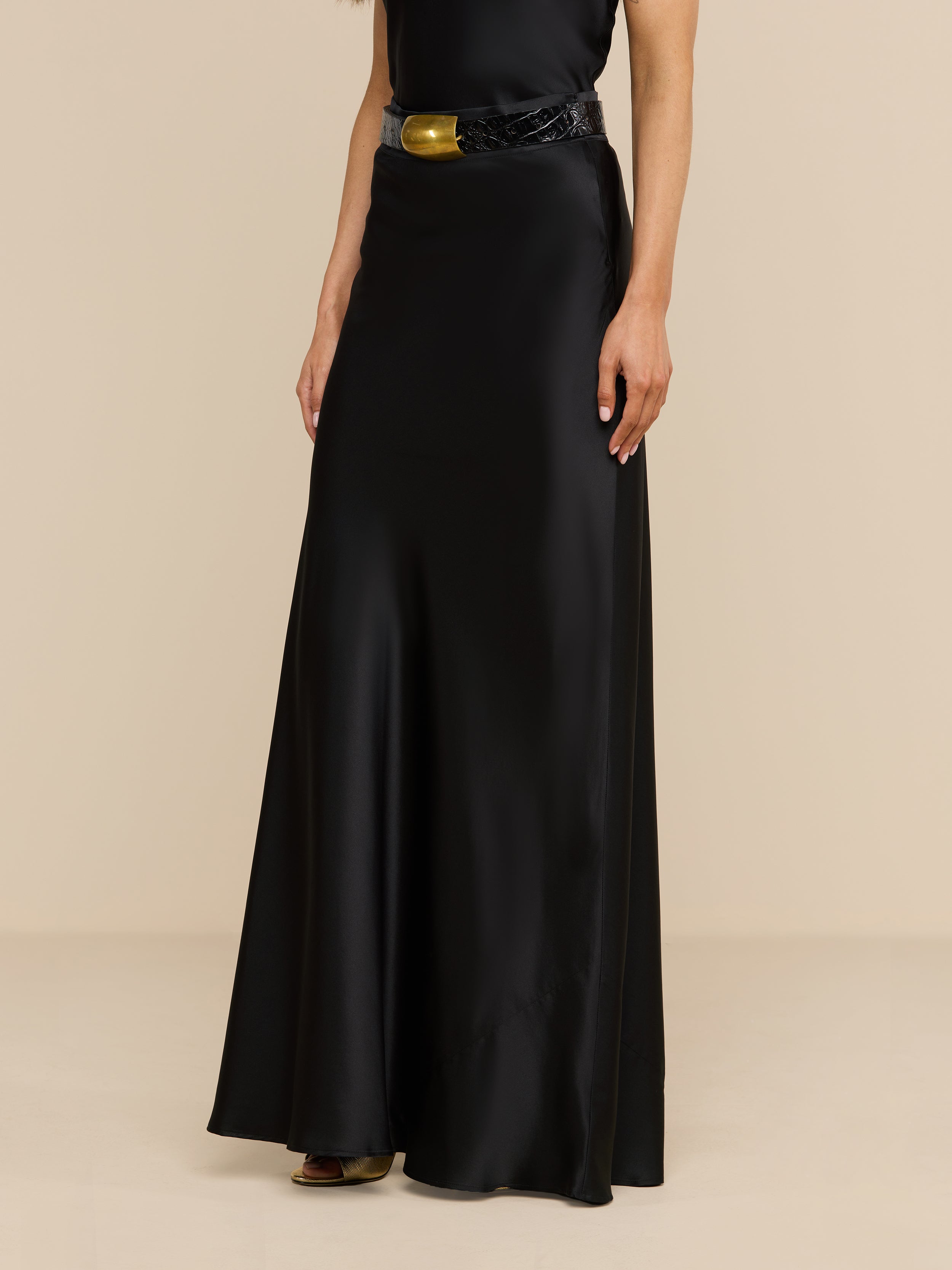 Wearing the Ryker Silk Maxi Skirt styled with a black belt featuring a bold gold buckle, the individual stands against a plain white background, embodying minimalist sophistication.