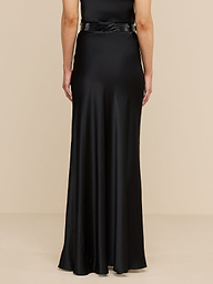 A person stands facing away, wearing the Ryker Silk Maxi Skirt in black, paired with a minimalist black belt for an effortlessly sophisticated look. The background is plain and white.