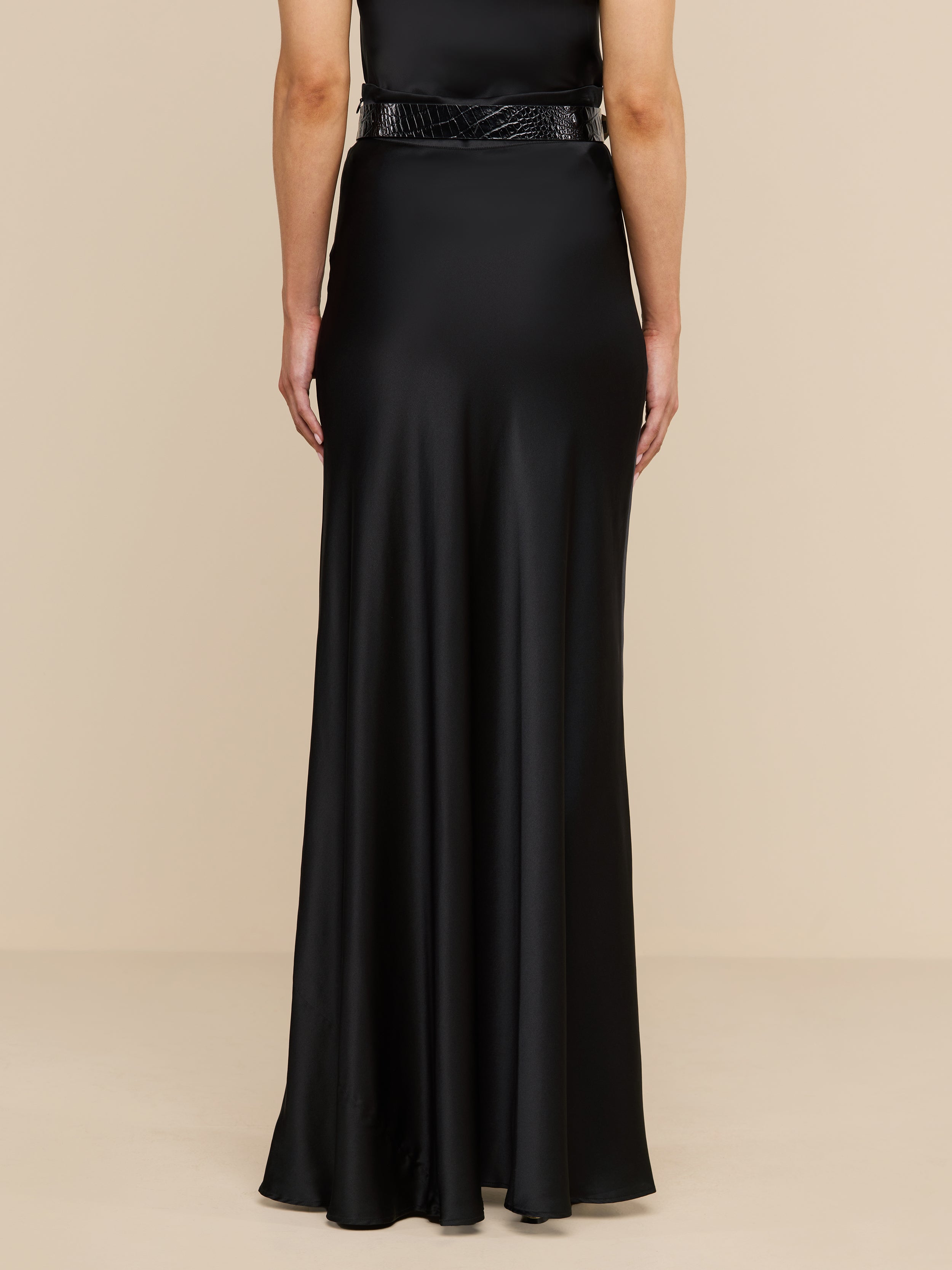 A person stands facing away, wearing the Ryker Silk Maxi Skirt in black, paired with a minimalist black belt for an effortlessly sophisticated look. The background is plain and white.