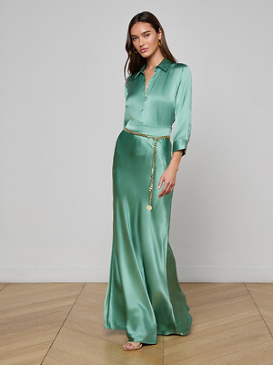 A woman in the Ryker Silk Maxi Skirt, a long jade green piece, stands on a wooden floor against a light wall. She wears open-toe heels, gold earrings, and a gold chain belt. Her long hair complements the skirt’s elegant look.