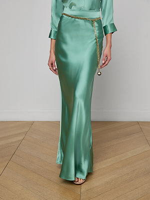 Someone is standing on a wooden floor wearing the Ryker Silk Maxi Skirt in shiny jade green, styled with a gold chain belt and a coordinating satin top. The person's face is not shown in the image.