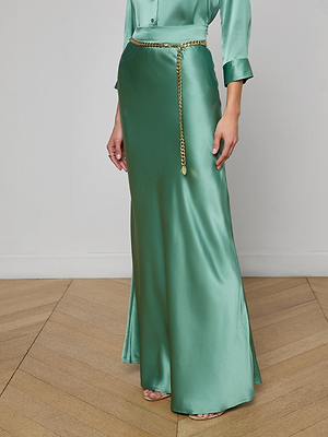 A person wearing the Ryker Silk Maxi Skirt in jade green with a matching shirt and thin gold chain belt, standing on a wooden floor against a plain white wall.