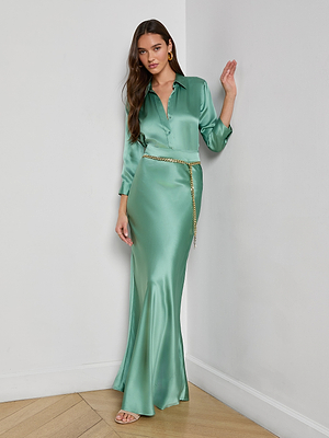 A woman stands indoors on a wooden floor, leaning against a white wall while wearing the Ryker Silk Maxi Skirt in jade green satin, styled with a gold chain belt.