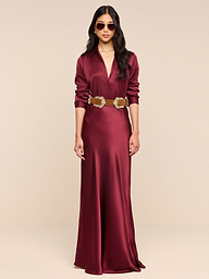 A woman stands against a plain white background, wearing the Ryker Silk Maxi Skirt in merlot red paired with a brown belt and sunglasses, looking forward with her hands by her sides.