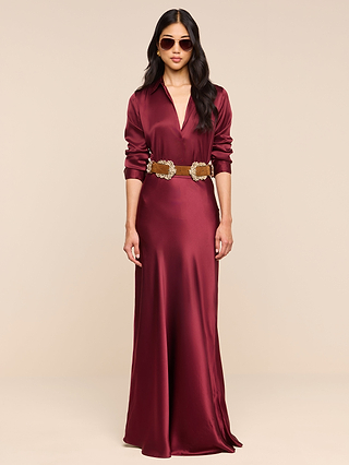 A woman pairs the Ryker Silk Maxi Skirt in merlot red with a deep V-neck top, wide brown belt featuring decorative buckles, chic sunglasses, and loose wavy hair, standing against a plain beige background.