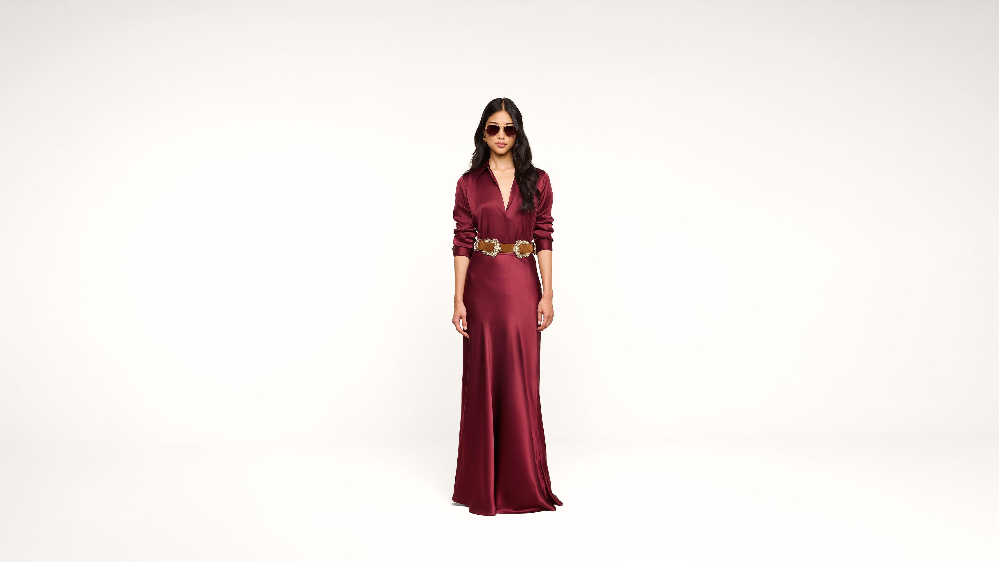 Woman stands in a long, deep red satin dress with a wide decorative belt and sunglasses against a plain white background.