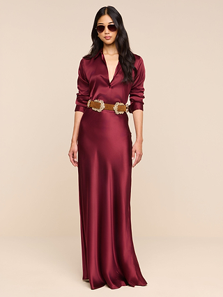 Wearing the Ryker Silk Maxi Skirt in a rich merlot red, this woman pairs its elegant, bias-cut drape with a deep-collared top, a brown belt featuring large buckles, statement sunglasses, and loose wavy hair. She stands poised against a plain beige background, her sophisticated style accentuated by the skirt’s luxurious silk finish.