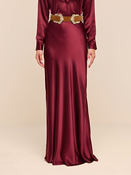A person wearing the Ryker Silk Maxi Skirt in a long, shiny merlot red hue, styled with long sleeves and a decorative gold belt and featuring a bias cut silhouette, stands against a plain white background.