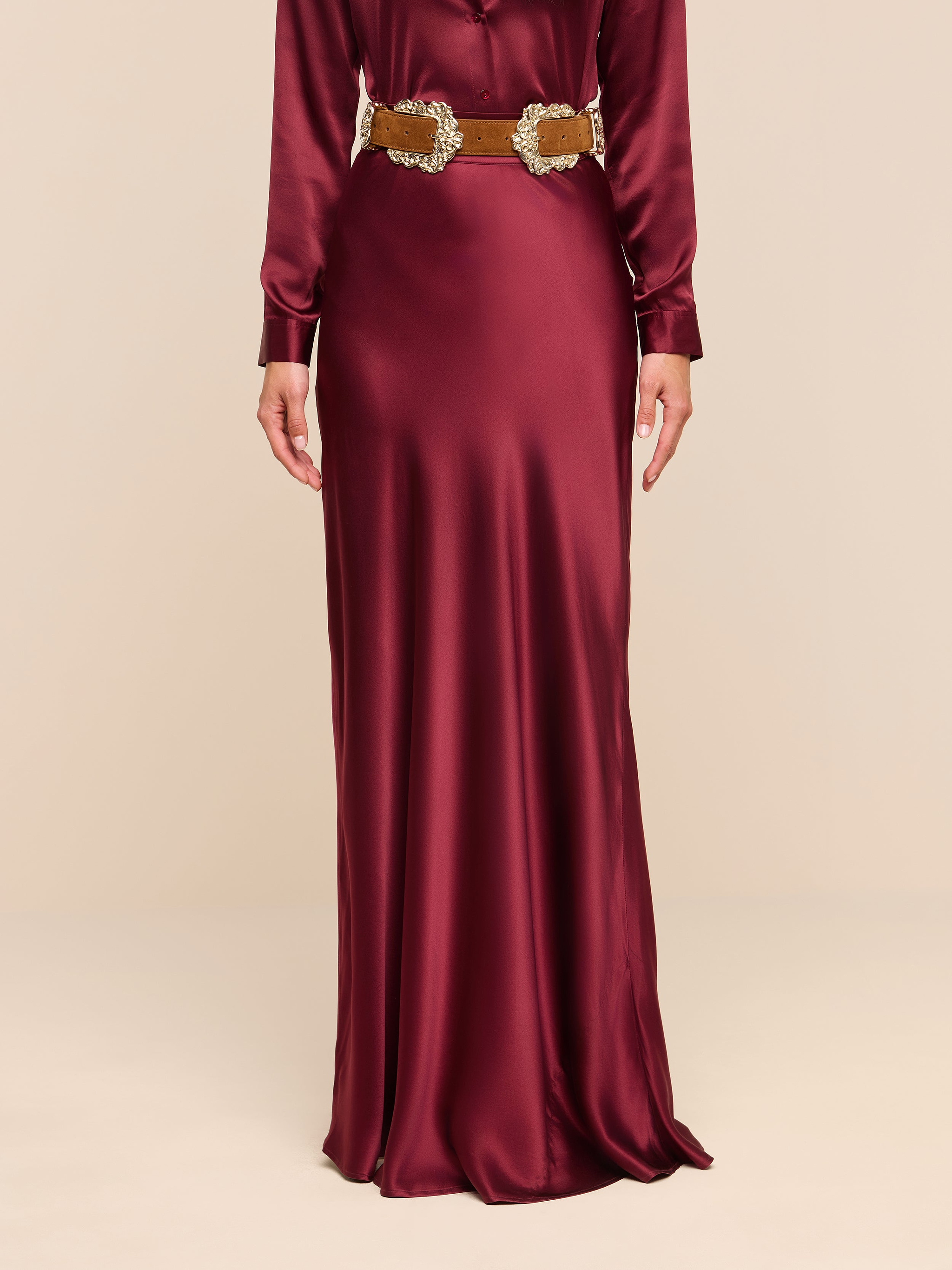A person wearing the Ryker Silk Maxi Skirt in a long, shiny merlot red hue, styled with long sleeves and a decorative gold belt and featuring a bias cut silhouette, stands against a plain white background.
