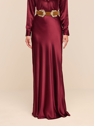 Wearing the Ryker Silk Maxi Skirt in a lustrous merlot red, paired with a wide tan belt adorned with oversized, ornate silver buckles, this look features an elegant bias-cut drape and long sleeves against a plain beige background.