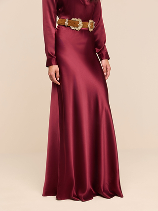 A person wearing the Ryker Silk Maxi Skirt in merlot red, styled with a brown belt featuring ornate gold details, stands against a plain beige background.