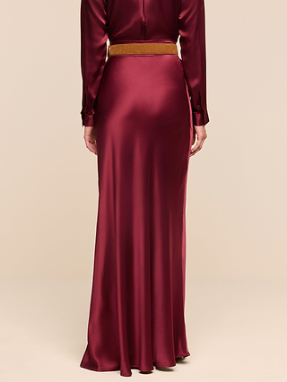 A person wearing the Ryker Silk Maxi Skirt in a long, merlot red hue with a brown belt, seen from the back against a plain beige background.