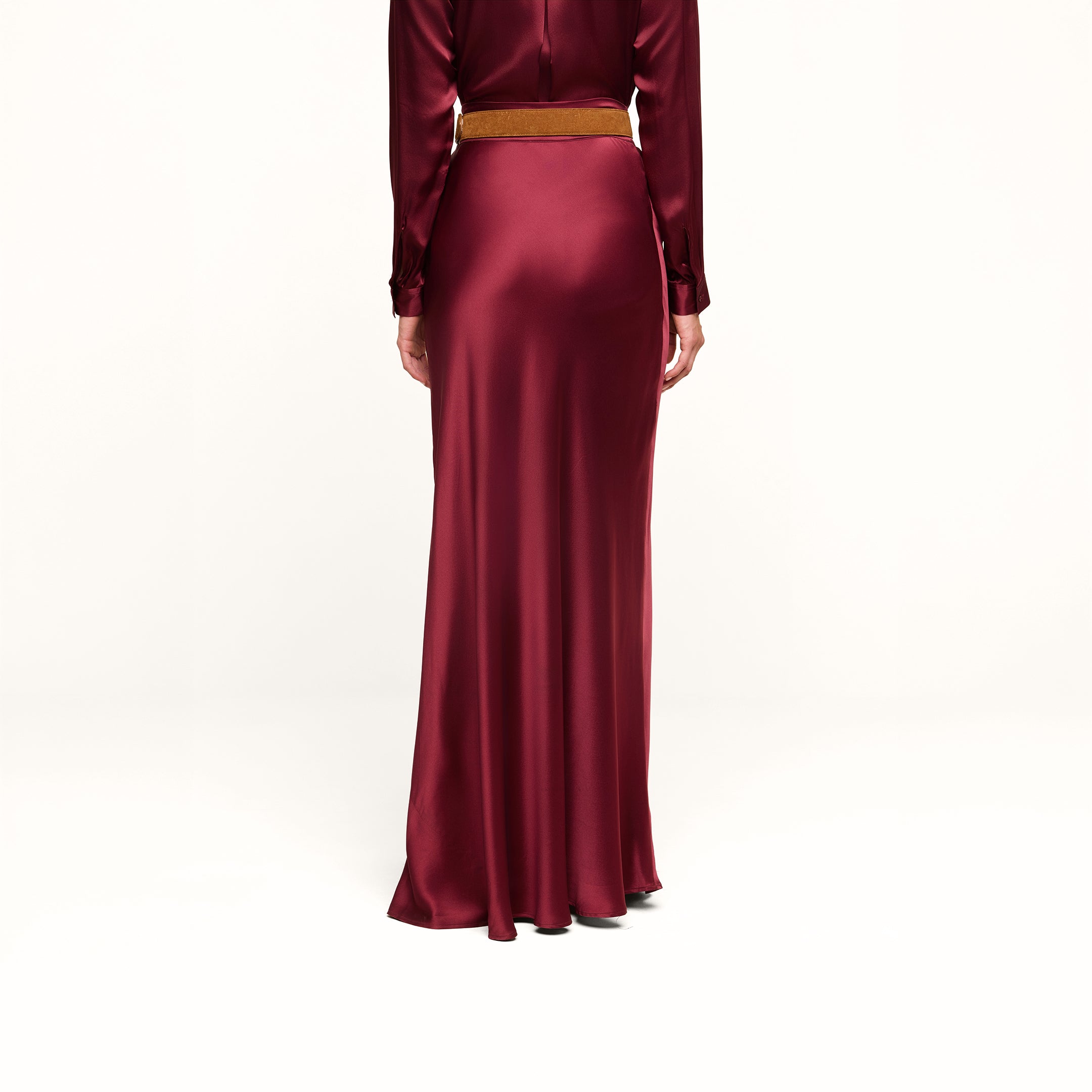 A person stands with their back to the camera, wearing the Ryker Silk Maxi Skirt in merlot red, styled with long sleeves and a brown belt, against a plain white background.