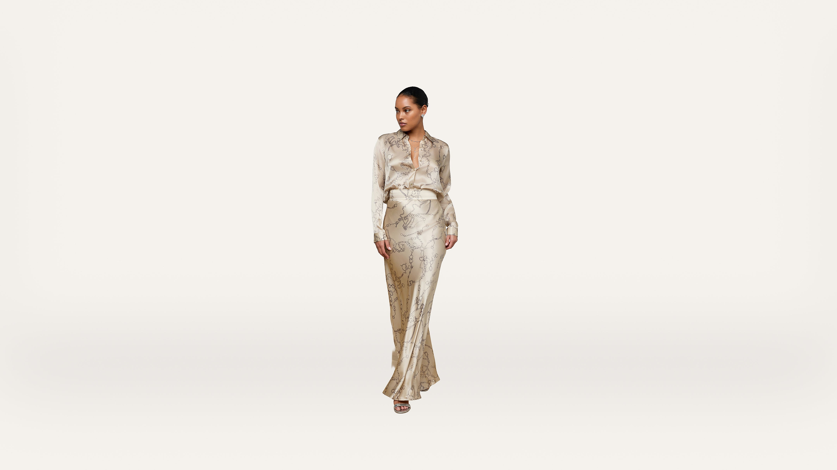 A woman stands confidently wearing a silky, light-colored, long-sleeve blouse and matching flowing skirt with a subtle pattern, against a plain, light background.