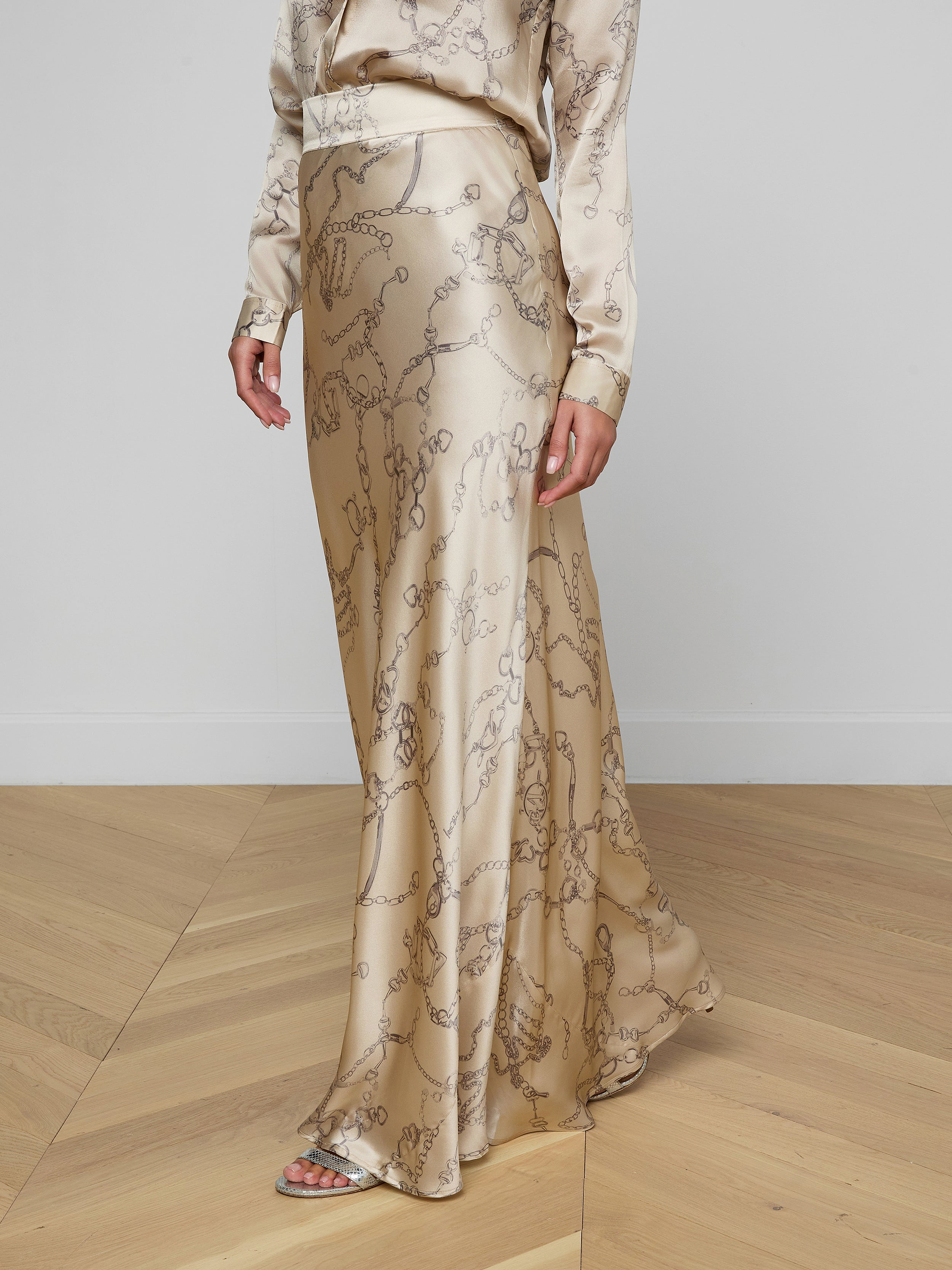 A person embodies minimalist sophistication in the Ryker Silk Maxi Skirt featuring a subtle horsebit chain print, paired with a matching blouse and open-toe sandals, standing on a light wood floor against a plain white wall.