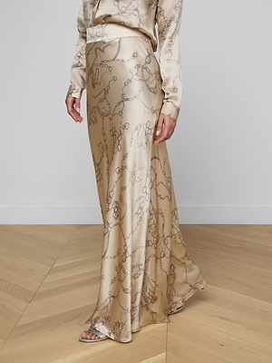 A person embodies minimalist sophistication in the Ryker Silk Maxi Skirt featuring a subtle horsebit chain print, paired with a matching blouse and open-toe sandals, standing on a light wood floor against a plain white wall.