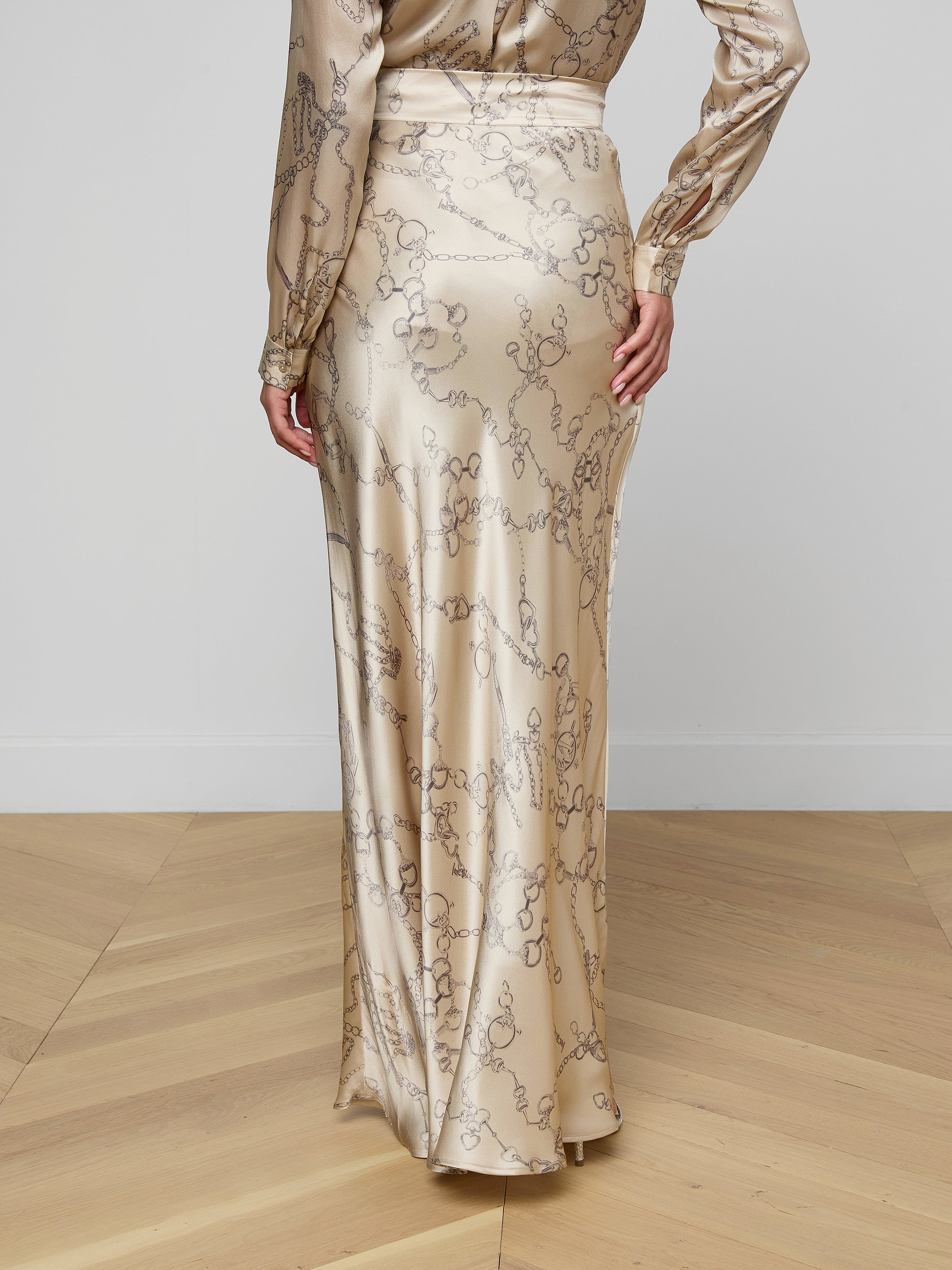 A person wearing the Ryker Silk Maxi Skirt, featuring a long, shiny design with a subtle abstract pattern, stands on a light wood floor facing away from the camera, showcasing minimalist sophistication.
