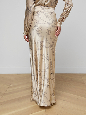 A person wearing the Ryker Silk Maxi Skirt, featuring a long, shiny design with a subtle abstract pattern, stands on a light wood floor facing away from the camera, showcasing minimalist sophistication.