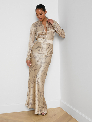 A woman exudes minimalist sophistication in a shiny, beige, long-sleeve blouse with the Ryker Silk Maxi Skirt featuring a horsebit chain print. She looks down, touching her collar and wears open-toed heels on a light wood floor.