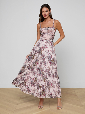 A woman stands on a wooden floor wearing the Torin Pleated Maxi Skirt in pink floral poly chiffon, styled with strappy heels. She holds part of the flared skirt, and the background is a plain white wall.