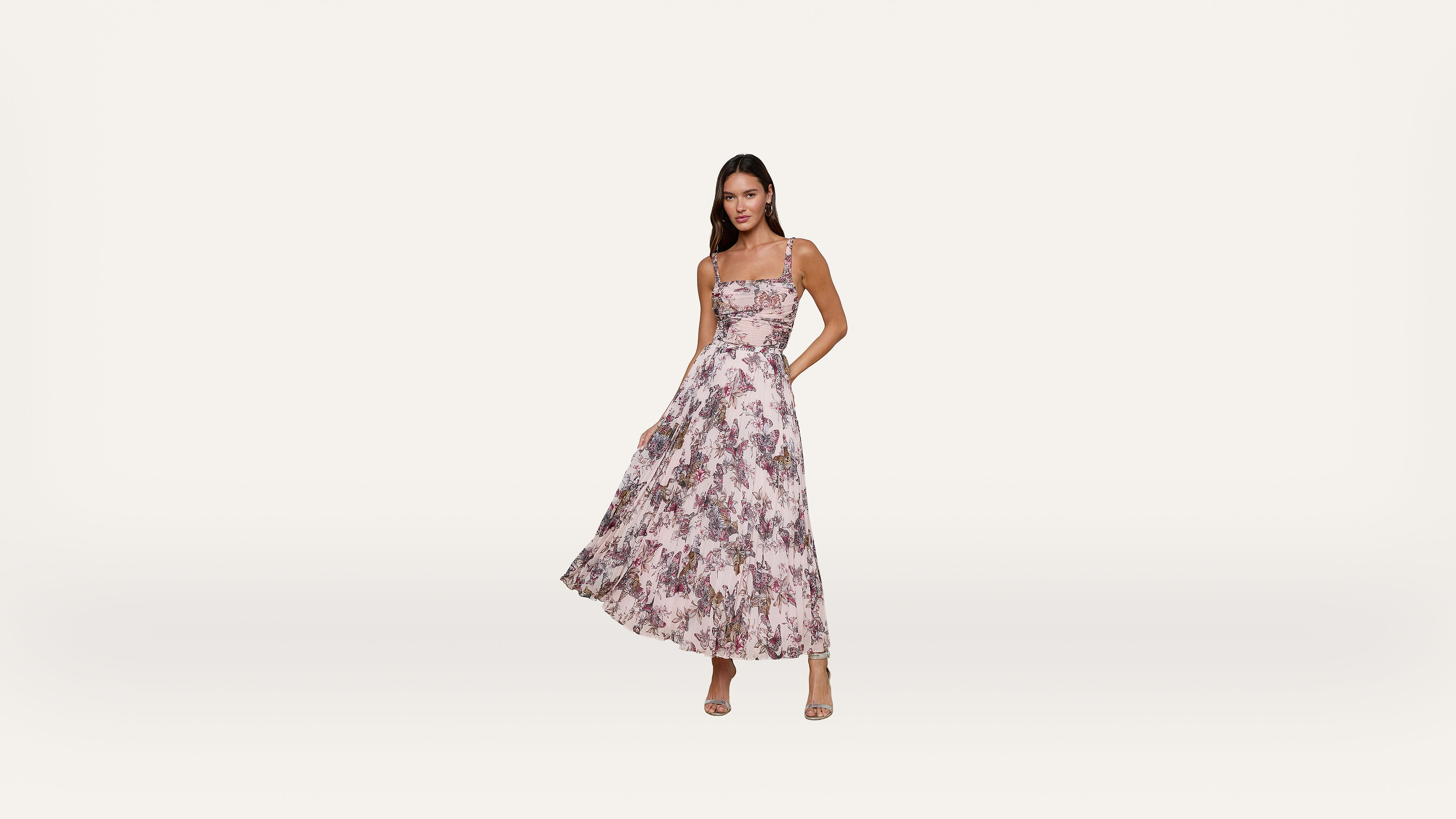 Woman in a sleeveless, pink and white floral dress with a flared skirt, posing with one hand on her hip against a plain light background.
