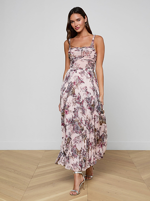 A woman poses indoors wearing the Torin Pleated Maxi Skirt in light pink poly chiffon with a floral print and flowing silhouette. She pairs it with strappy heeled sandals and has long hair.