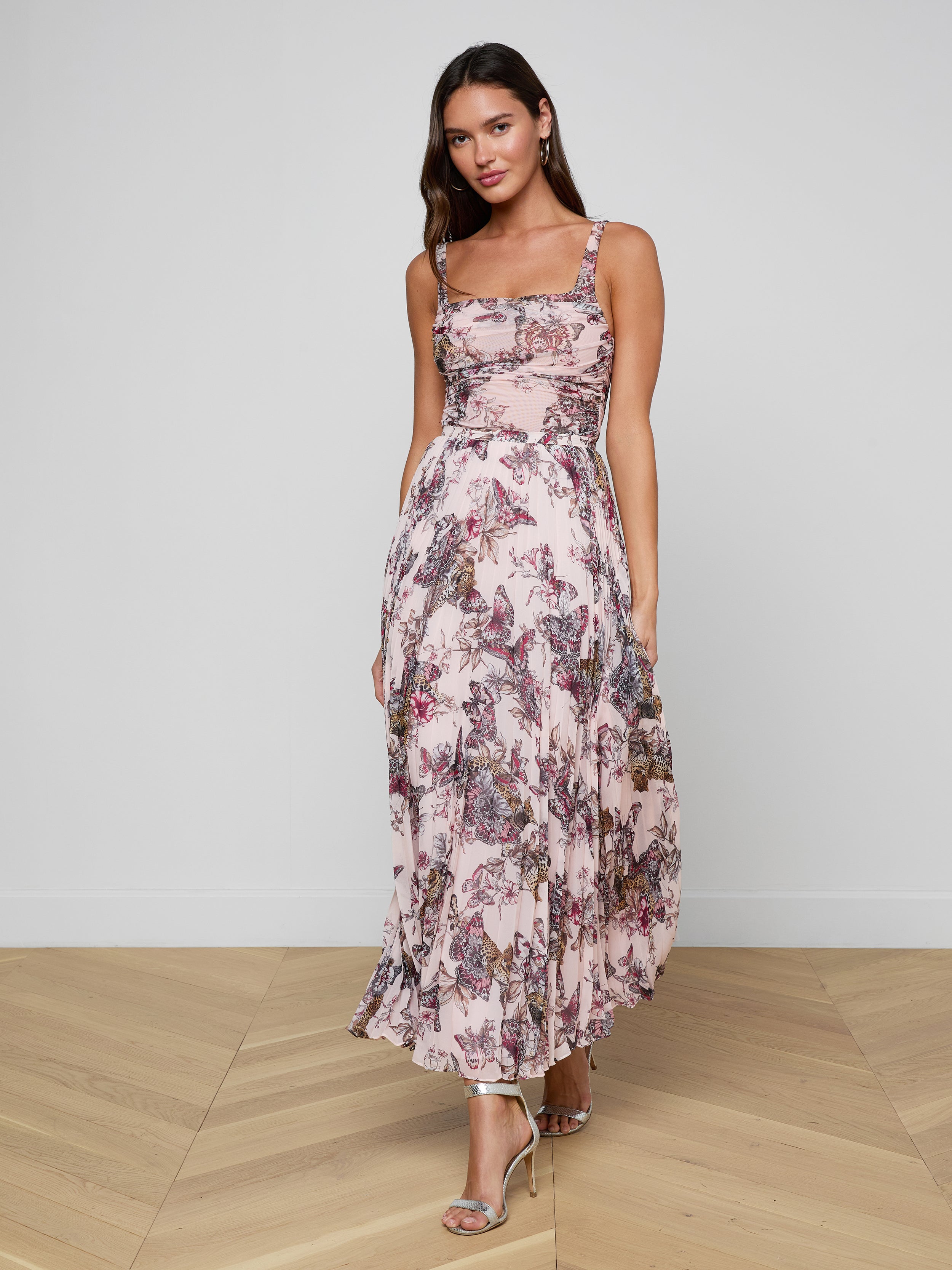 A woman stands against a plain white background, wearing the Torin Pleated Maxi Skirt—a pink floral poly chiffon maxi skirt with elegant pleats—styled with a sleeveless fitted bodice and strappy high-heeled sandals.