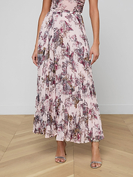 A woman wearing the Torin Pleated Maxi Skirt, styled with a sleeveless, pink floral poly chiffon maxi dress and open-toe heels, stands against a plain white background. Her head and feet are not visible in the image.