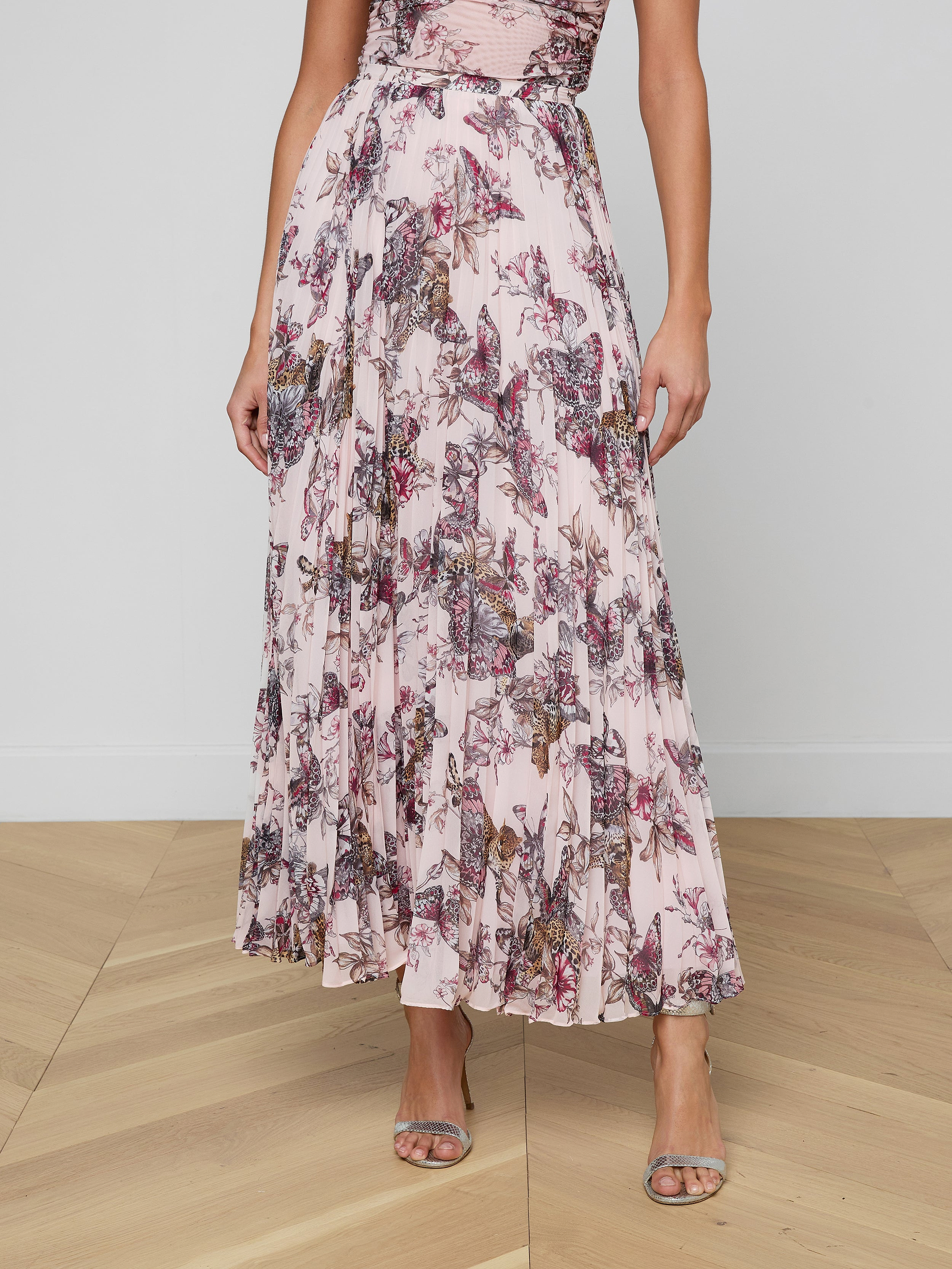 A woman wearing the Torin Pleated Maxi Skirt, styled with a sleeveless, pink floral poly chiffon maxi dress and open-toe heels, stands against a plain white background. Her head and feet are not visible in the image.