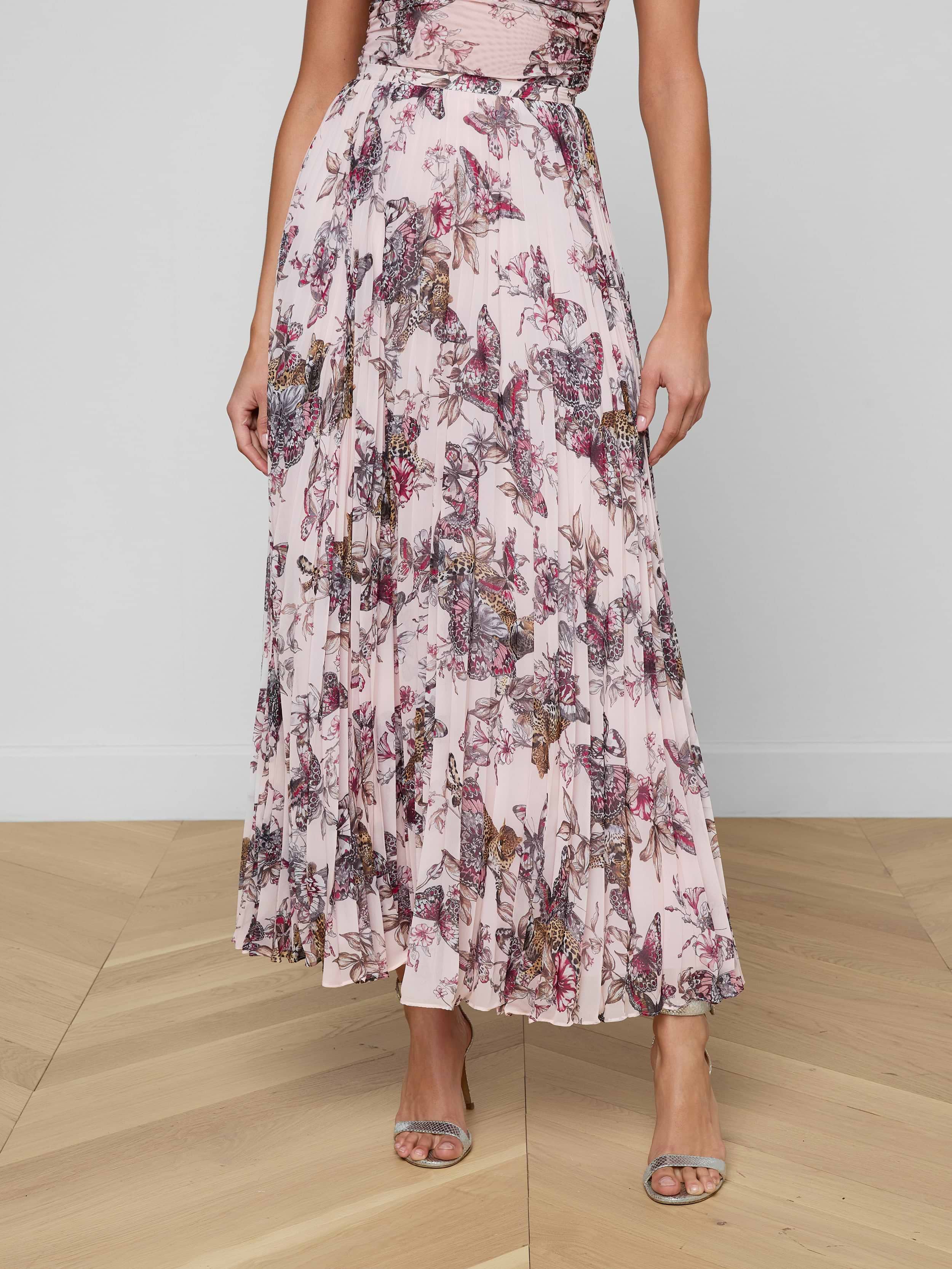 A woman wearing the Torin Pleated Maxi Skirt, styled with a sleeveless, pink floral poly chiffon maxi dress and open-toe heels, stands against a plain white background. Her head and feet are not visible in the image.