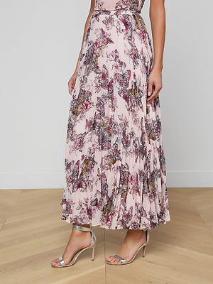 A woman stands on a light wood floor against a white wall, wearing the Torin Pleated Maxi Skirt in light pink poly chiffon with floral, butterfly, and jungle print details, paired with silver high-heeled sandals.