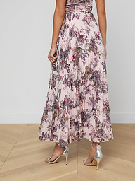 A woman shown from the back on a plain light background wears the Torin Pleated Maxi Skirt in a flowing, ankle-length, pink floral poly chiffon fabric, paired with silver high-heeled sandals.