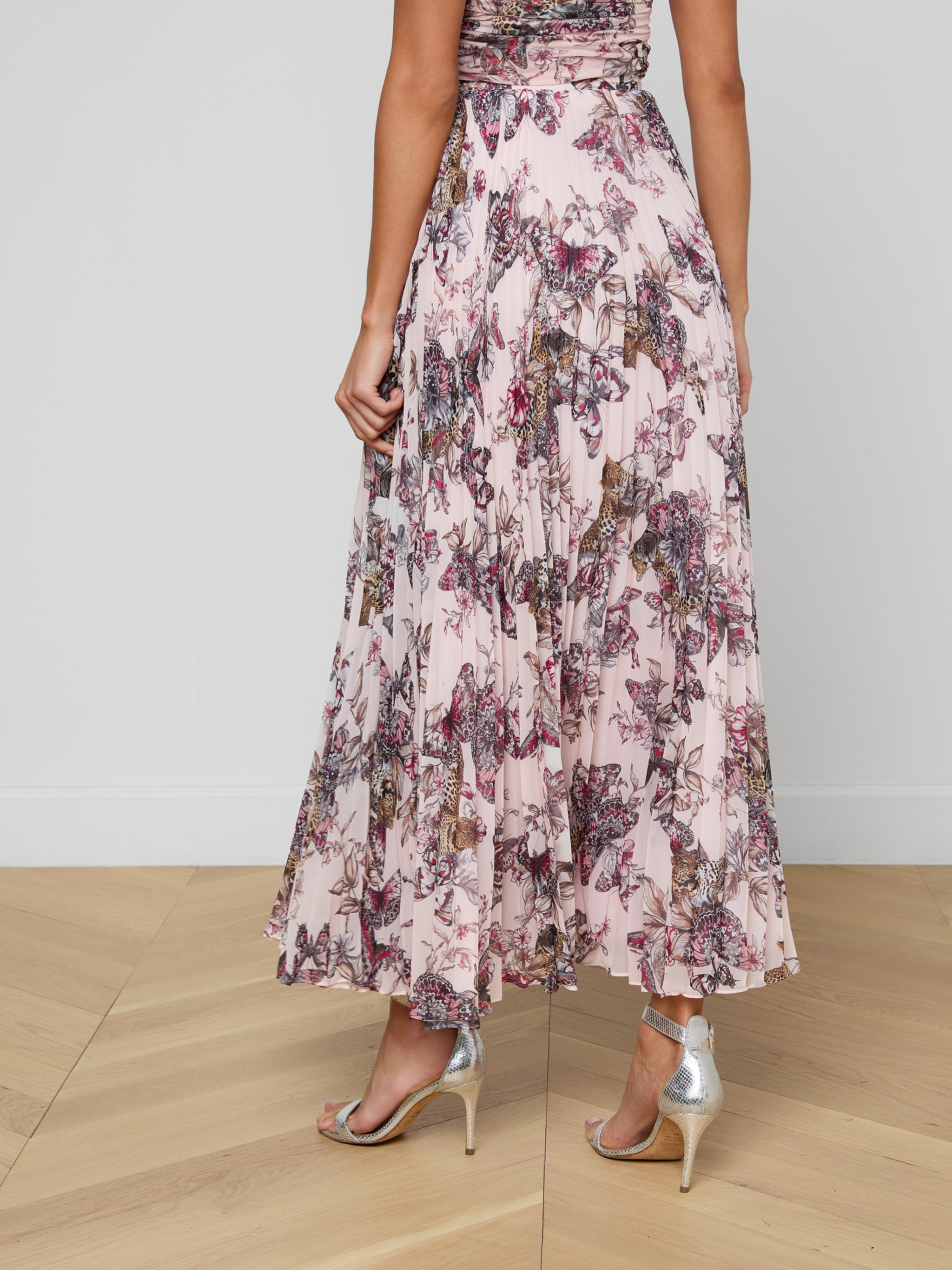 A woman shown from the back on a plain light background wears the Torin Pleated Maxi Skirt in a flowing, ankle-length, pink floral poly chiffon fabric, paired with silver high-heeled sandals.