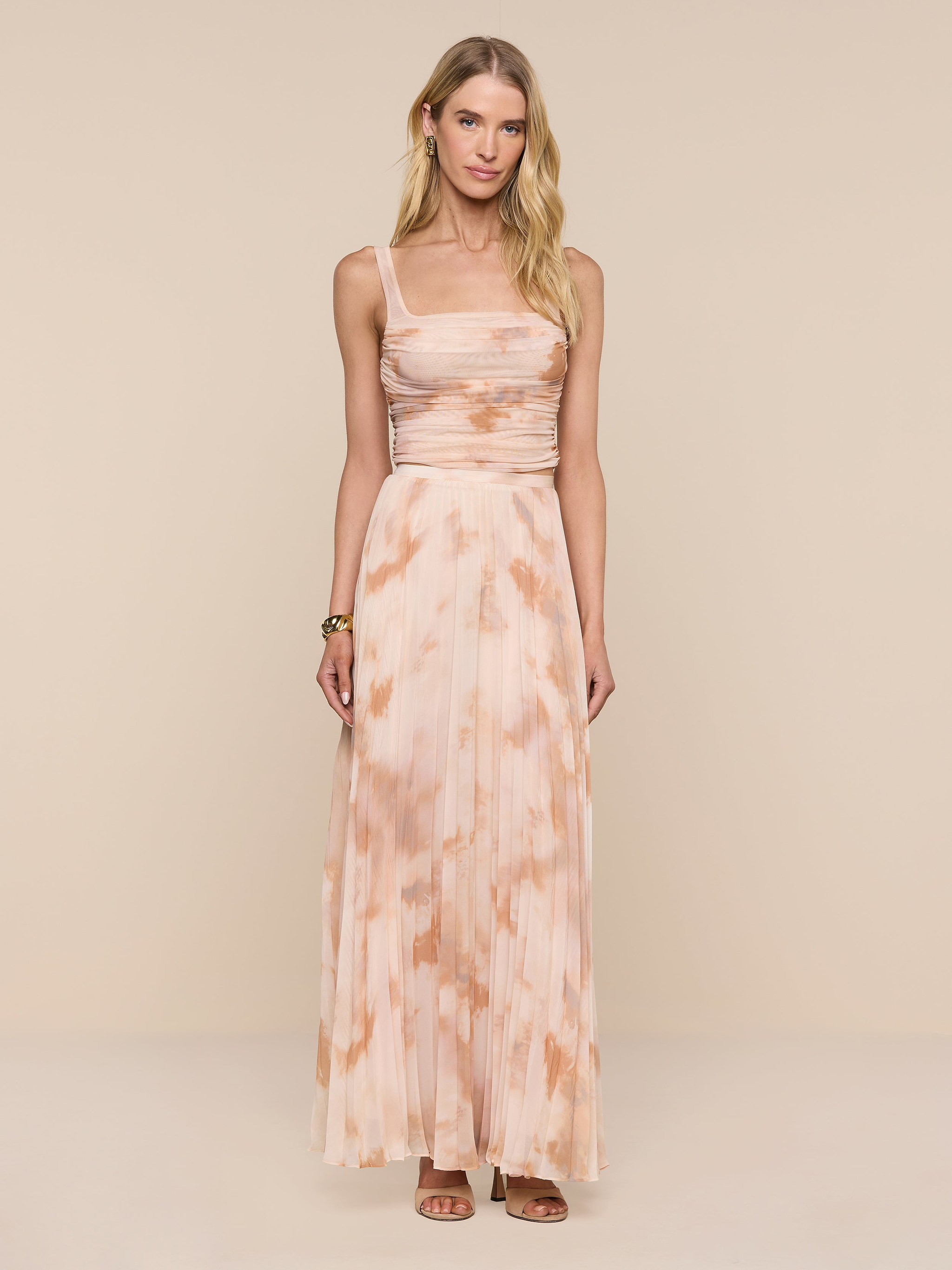 A woman stands against a plain white background, wearing the Torin Pleated Maxi Skirt in a long, peach and beige tie-dye design. She pairs the airy poly chiffon skirt with a sleeveless top featuring thin straps and a ruched bodice. Her long blonde hair flows down, and she accessorizes with brown sandals and a bracelet.