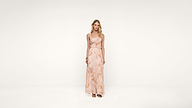 A woman stands in a light pink, sleeveless, floor-length dress with a subtle pattern, against a plain white background. She has long blonde hair and wears sandals.