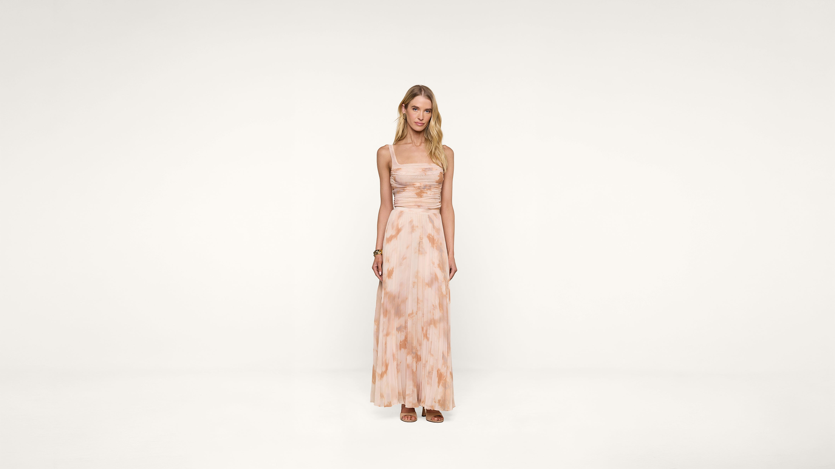 A woman stands in a light pink, sleeveless, floor-length dress with a subtle pattern, against a plain white background. She has long blonde hair and wears sandals.